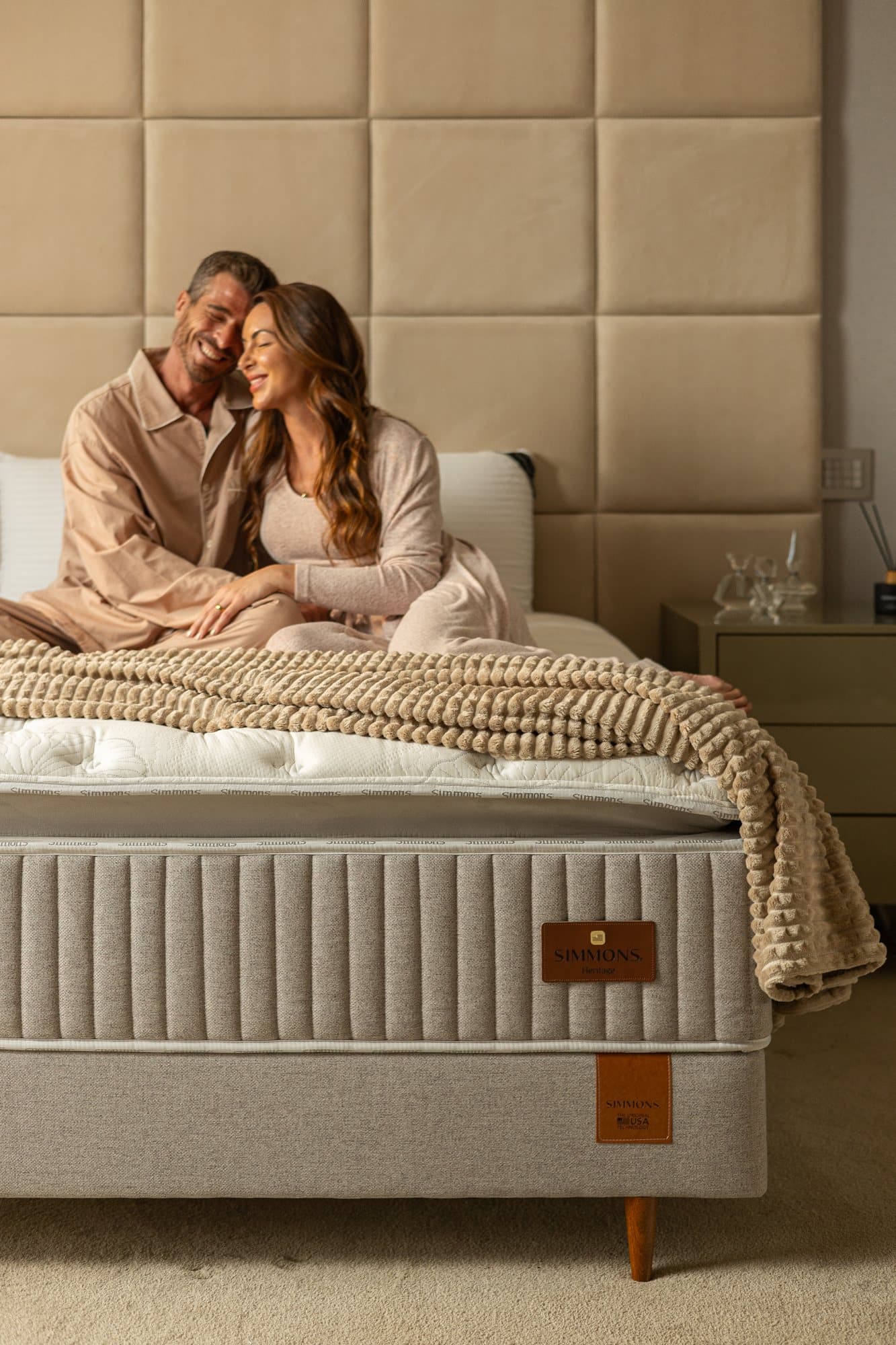Casal de pijama abraçado e rindo, sentado na cama com cabeceira estofada alta e colchão Simmons Heritage